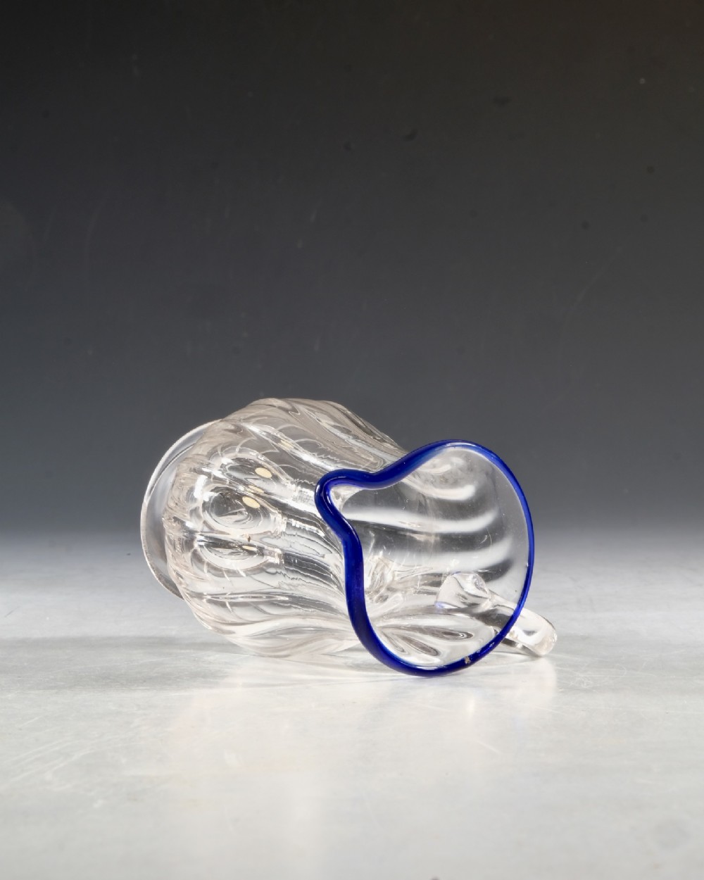 An English Georgian blown clear glass cream jug featuring vertical fluting, a loop handle, and an applied cobalt blue rim. View V