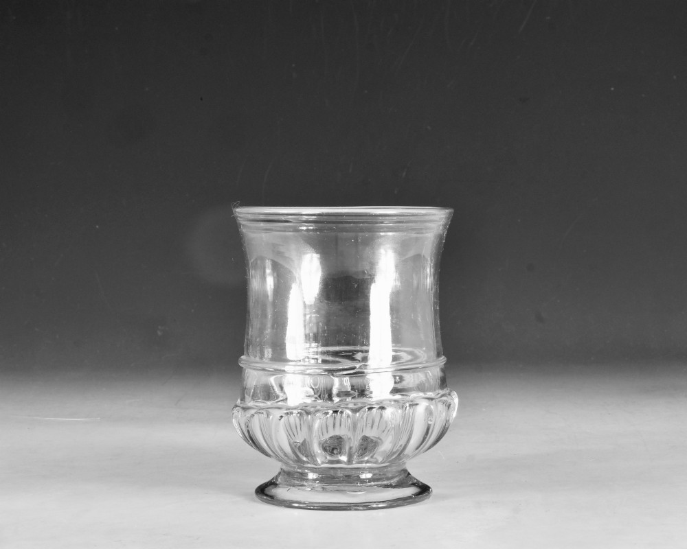 A clear 18th-century Georgian glass drinking vessel featuring a flared rim, a ribbed lower body, and a low circular pedestal base.