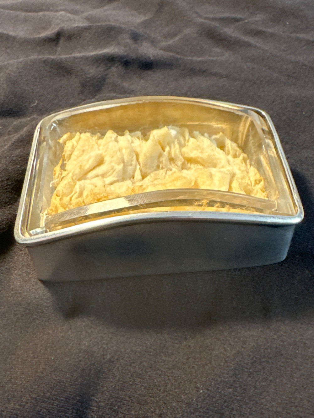 A rectangular silver vessel with a curved rim containing a light yellow natural sponge.