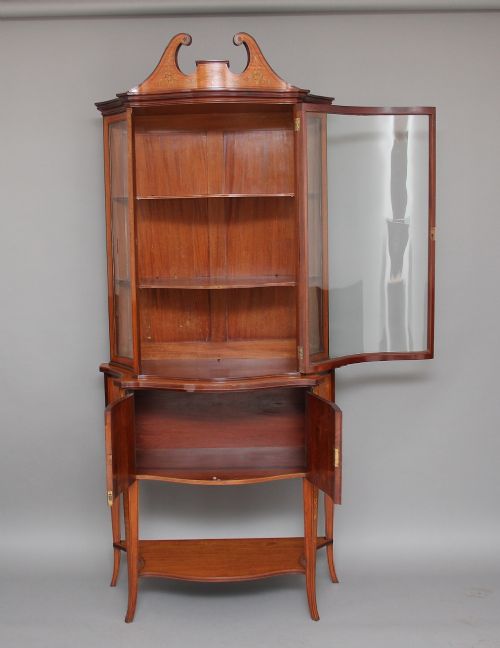 A 19th-century satinwood display cabinet featuring a swan-neck pediment, curved central glass door, and marquetry inlaid lower doors on square tapered legs. View II