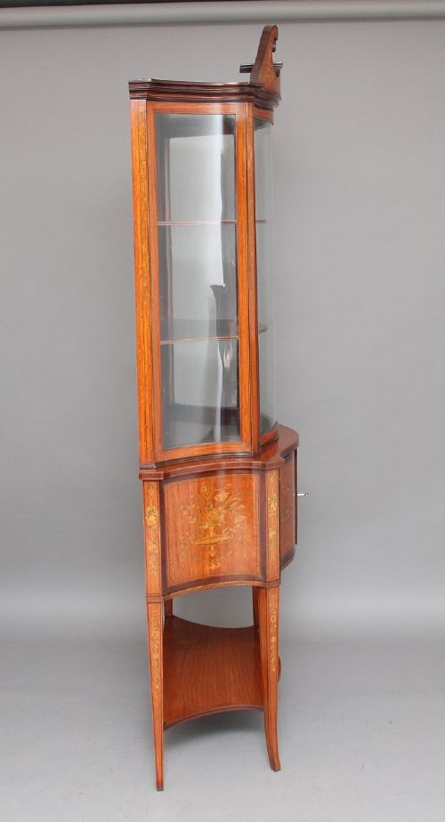 A 19th-century satinwood display cabinet featuring a swan-neck pediment, curved central glass door, and marquetry inlaid lower doors on square tapered legs. View V