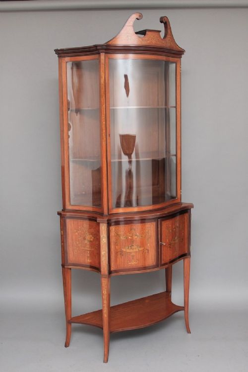 A 19th-century satinwood display cabinet featuring a swan-neck pediment, curved central glass door, and marquetry inlaid lower doors on square tapered legs. View VII