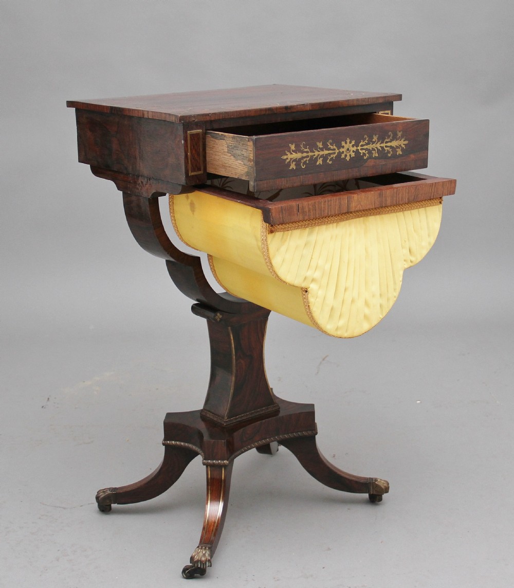 An early 19th-century dark rosewood sewing table featuring brass marquetry inlay on the drawer front, a yellow pleated fabric work basket, and a pedestal base on quadform sabre legs with brass feet. View IV