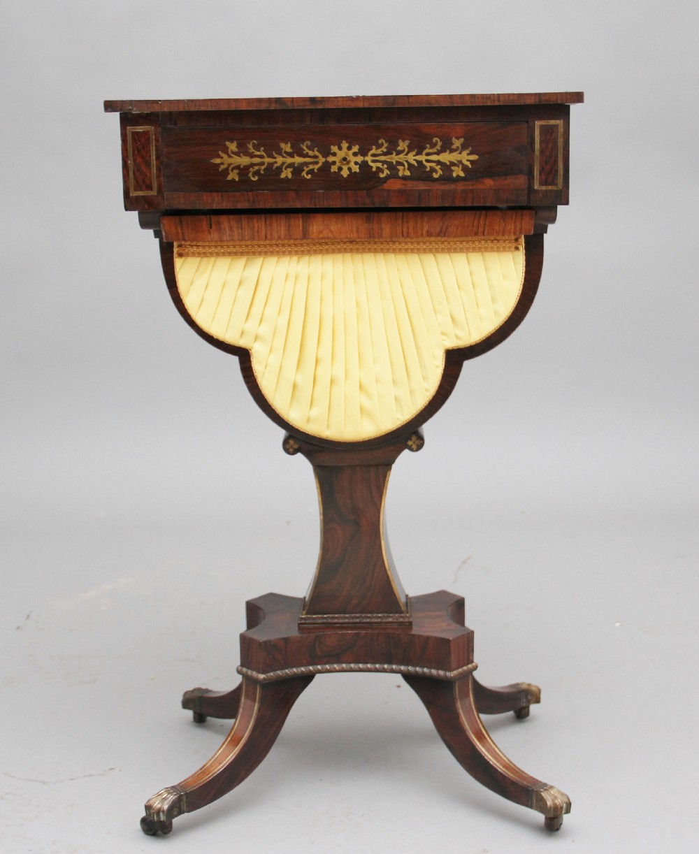 An early 19th-century dark rosewood sewing table featuring brass marquetry inlay on the drawer front, a yellow pleated fabric work basket, and a pedestal base on quadform sabre legs with brass feet. View V