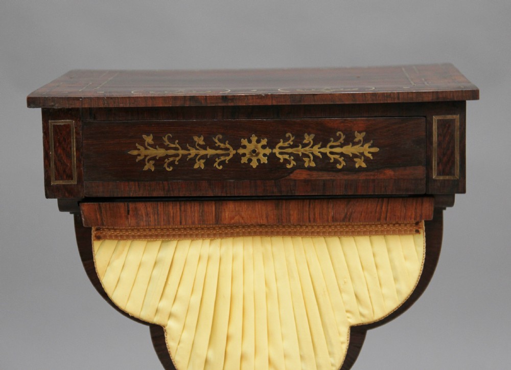 An early 19th-century dark rosewood sewing table featuring brass marquetry inlay on the drawer front, a yellow pleated fabric work basket, and a pedestal base on quadform sabre legs with brass feet. View X