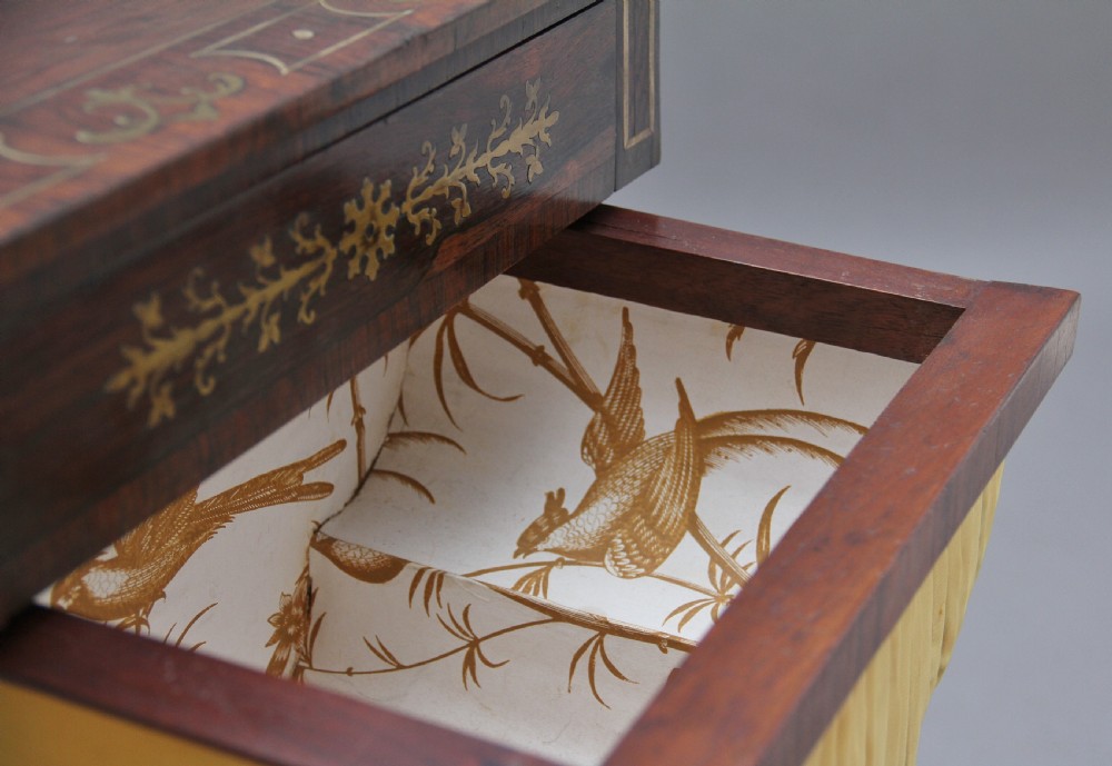 An early 19th-century dark rosewood sewing table featuring brass marquetry inlay on the drawer front, a yellow pleated fabric work basket, and a pedestal base on quadform sabre legs with brass feet. View XIII