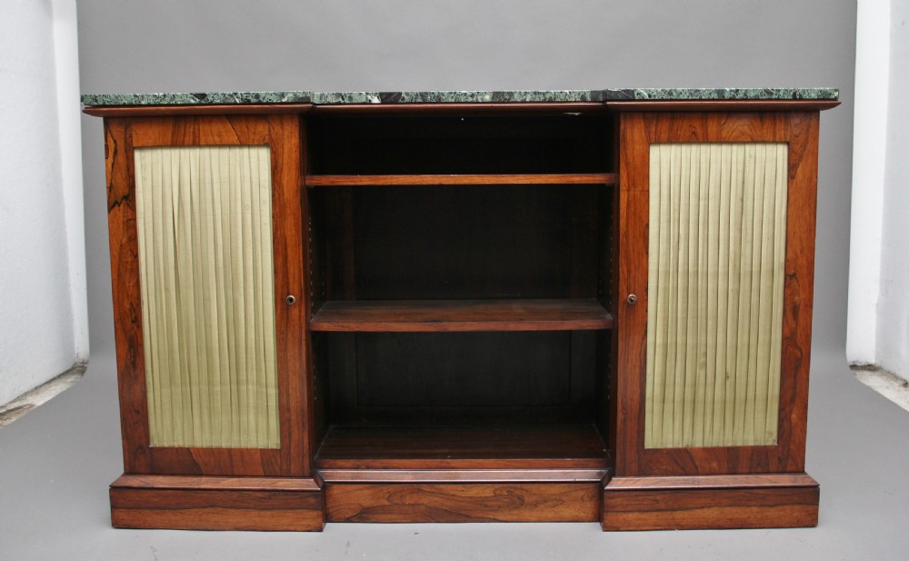 An early nineteenth-century rosewood breakfront cabinet featuring a green marble top, open central shelving section, and flanking doors with pleated fabric panels on a solid plinth base.