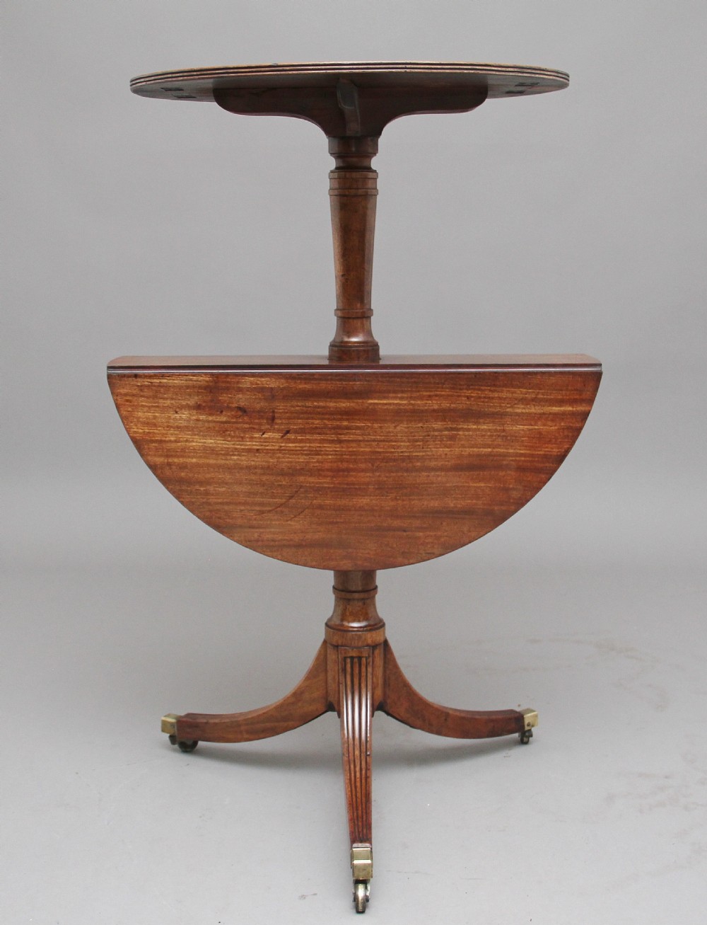 An early nineteenth-century mahogany two-tier dumbwaiter featuring circular drop-leaf surfaces, a turned central column support, and reeded tripod legs with brass castors. View VII