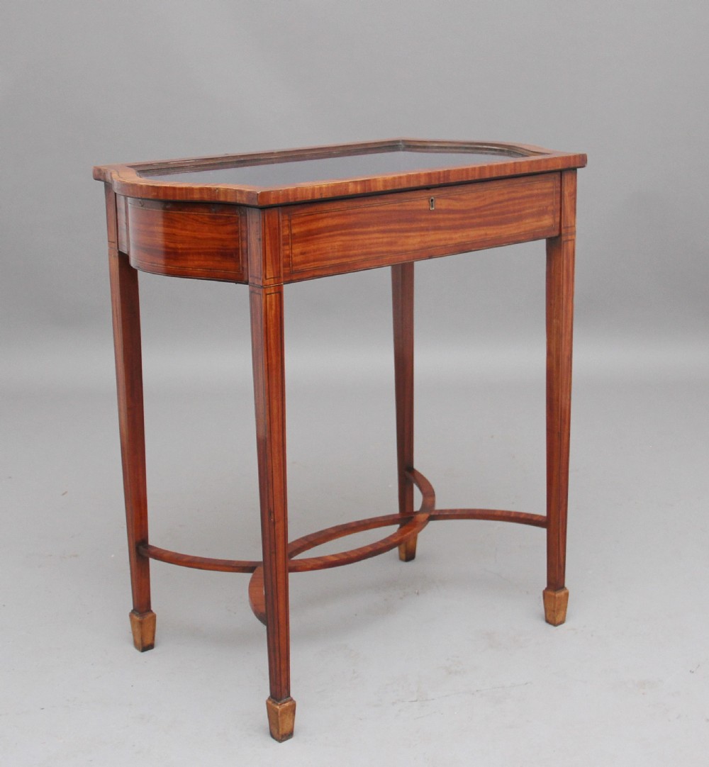 An Edwardian inlaid satinwood bijouterie table featuring a glazed rectangular top, curved side profile, tapered square legs, and curved cross-stretchers.