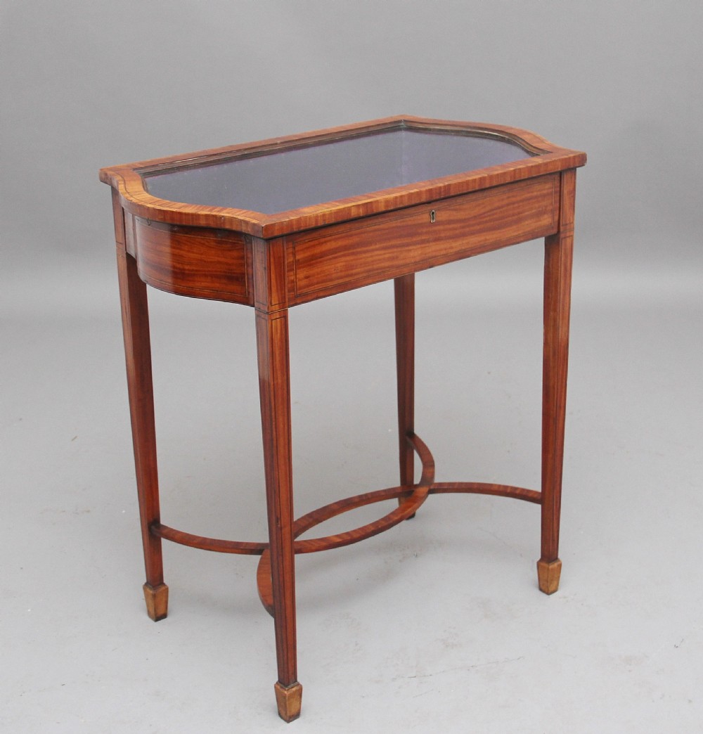 An Edwardian inlaid satinwood bijouterie table featuring a glazed rectangular top, curved side profile, tapered square legs, and curved cross-stretchers. View IV