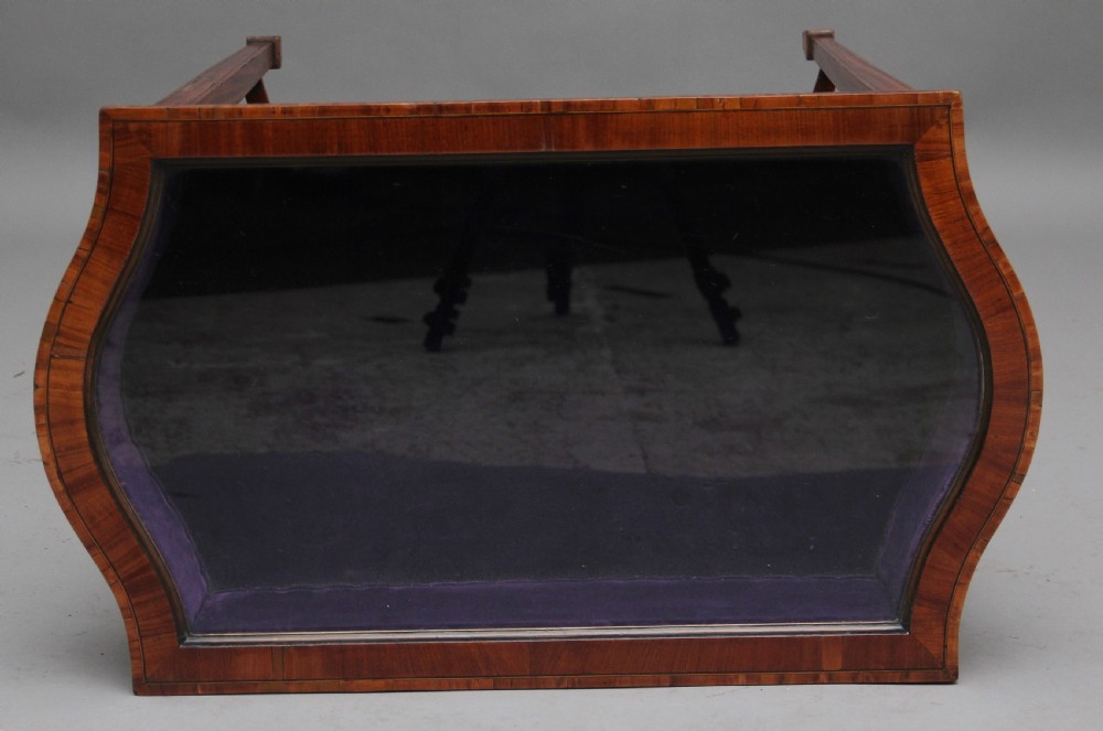 An Edwardian inlaid satinwood bijouterie table featuring a glazed rectangular top, curved side profile, tapered square legs, and curved cross-stretchers. View V
