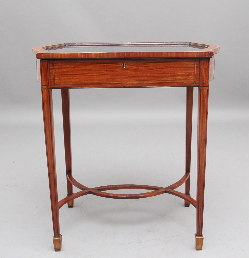 An Edwardian inlaid satinwood bijouterie table featuring a glazed rectangular top, curved side profile, tapered square legs, and curved cross-stretchers. View VI