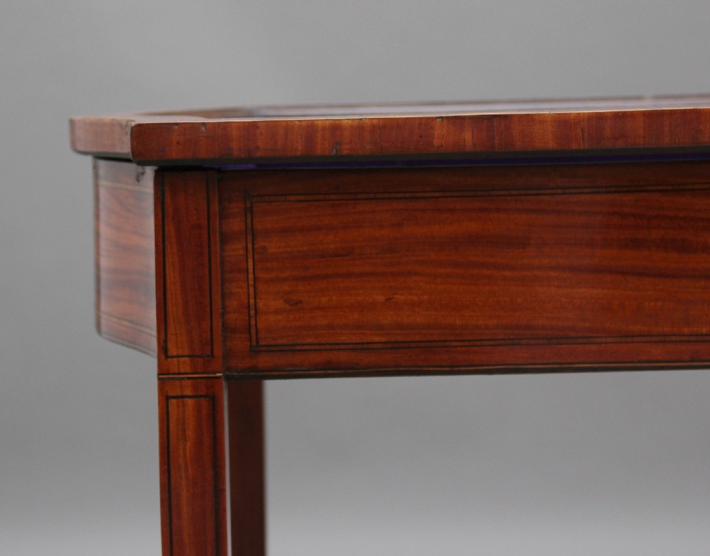 An Edwardian inlaid satinwood bijouterie table featuring a glazed rectangular top, curved side profile, tapered square legs, and curved cross-stretchers. View X