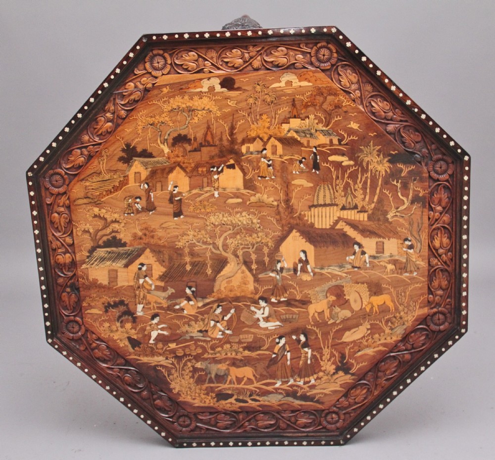 An octagonal wooden coffee table featuring detailed pictorial wood marquetry depicting an Indian village scene surrounded by a relief-carved floral border and ebonised outer rim.