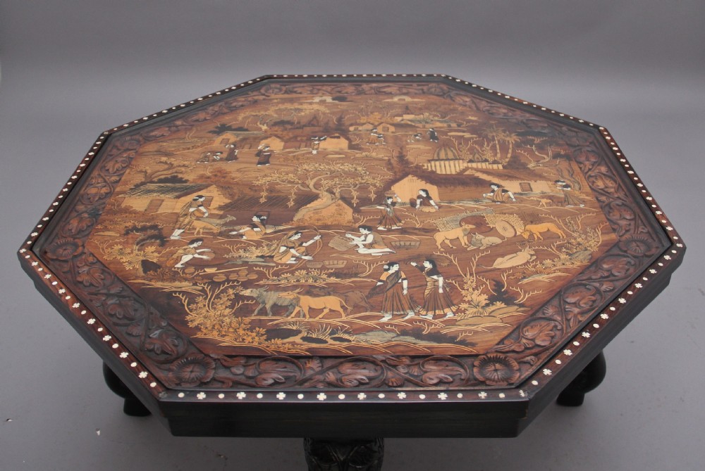 An octagonal wooden coffee table featuring detailed pictorial wood marquetry depicting an Indian village scene surrounded by a relief-carved floral border and ebonised outer rim. View VIII