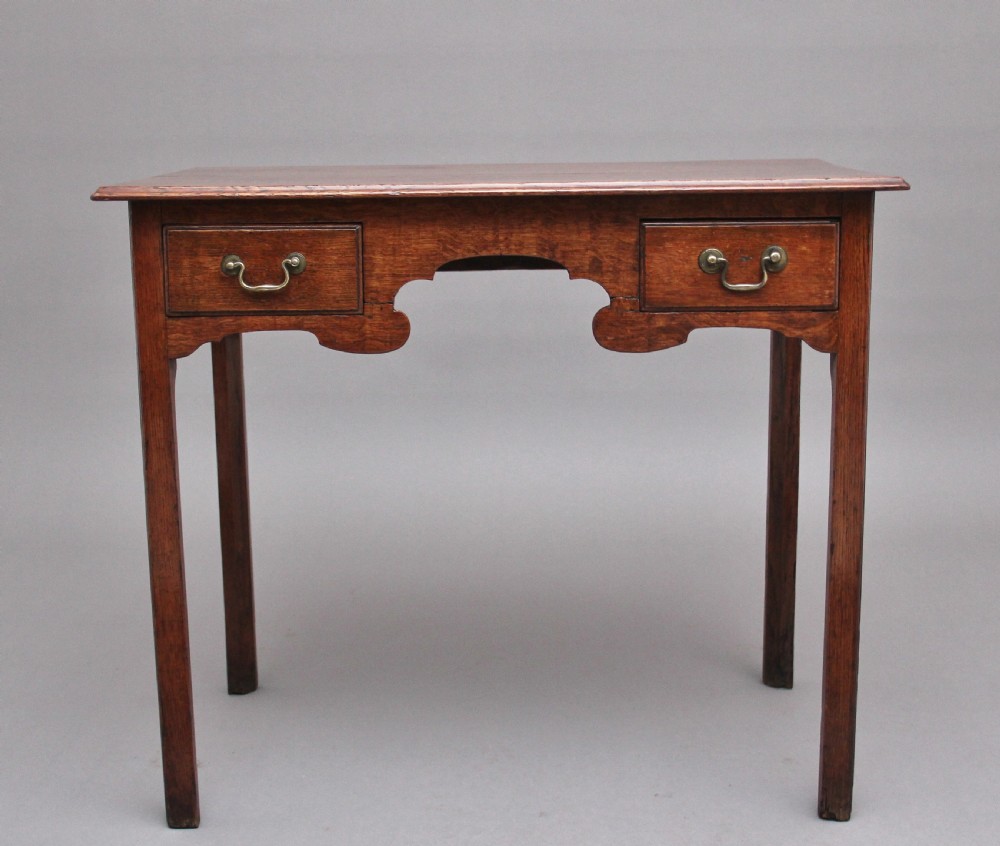 An early 19th-century oak lowboy side table featuring two drawers with brass handles, a shaped lower apron, and four straight legs.