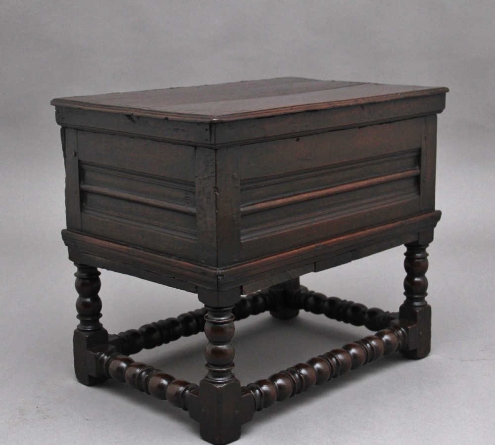 An early 18th-century dark oak box stool featuring a hinged lid, moulded side panels, and turned legs joined by stretchers.