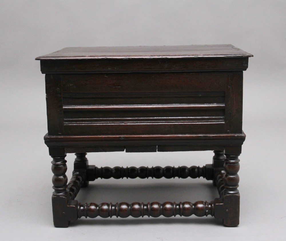 An early 18th-century dark oak box stool featuring a hinged lid, moulded side panels, and turned legs joined by stretchers. View II