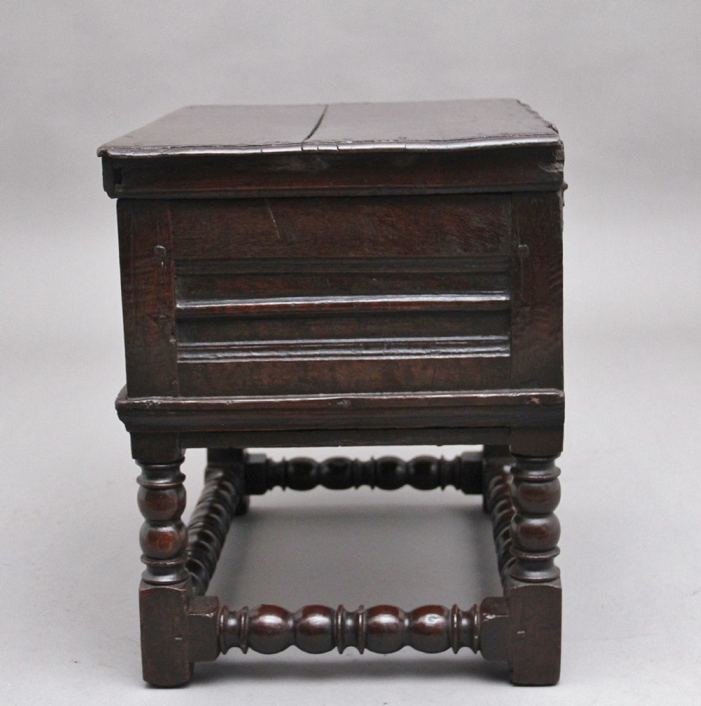 An early 18th-century dark oak box stool featuring a hinged lid, moulded side panels, and turned legs joined by stretchers. View V