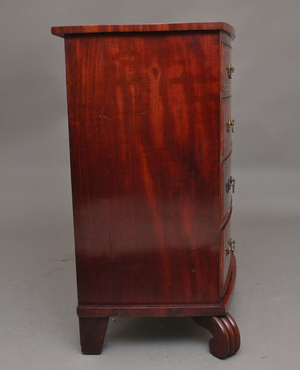 An early nineteenth-century flame mahogany bowfront chest featuring four graduated drawers with inlaid banding and brass handles. View V