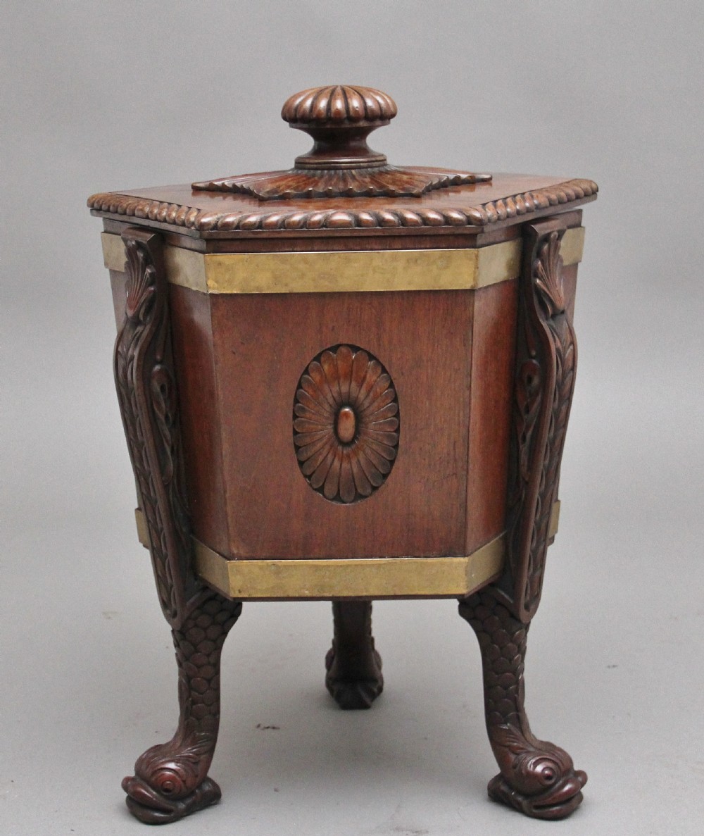 An early twentieth-century mahogany wine cooler featuring a gadrooned hinged lid with turned finial, brass strapping, carved rosette details, and carved dolphin legs.