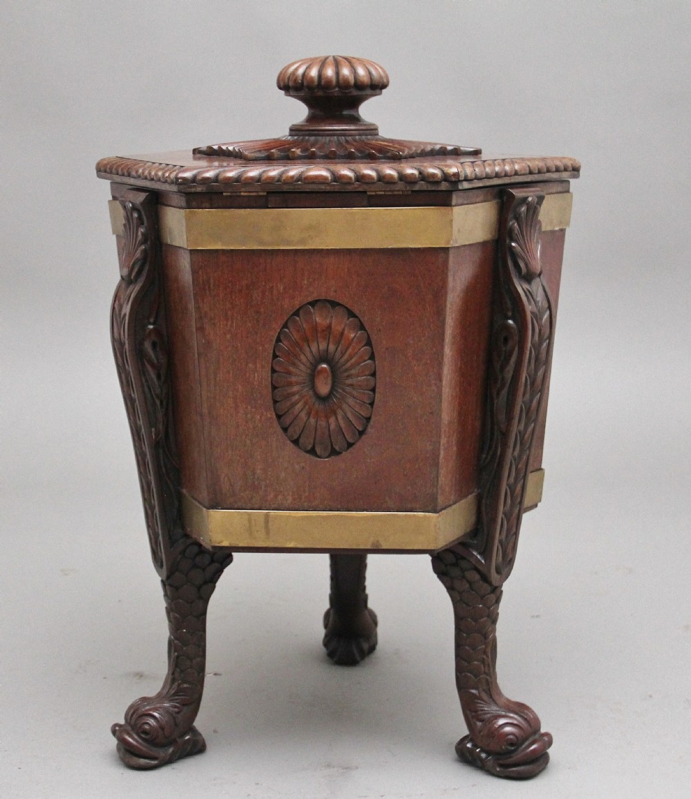 An early twentieth-century mahogany wine cooler featuring a gadrooned hinged lid with turned finial, brass strapping, carved rosette details, and carved dolphin legs. View V
