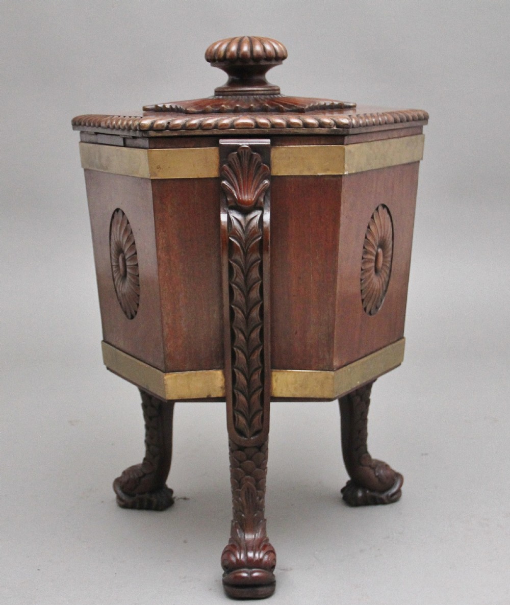 An early twentieth-century mahogany wine cooler featuring a gadrooned hinged lid with turned finial, brass strapping, carved rosette details, and carved dolphin legs. View VI
