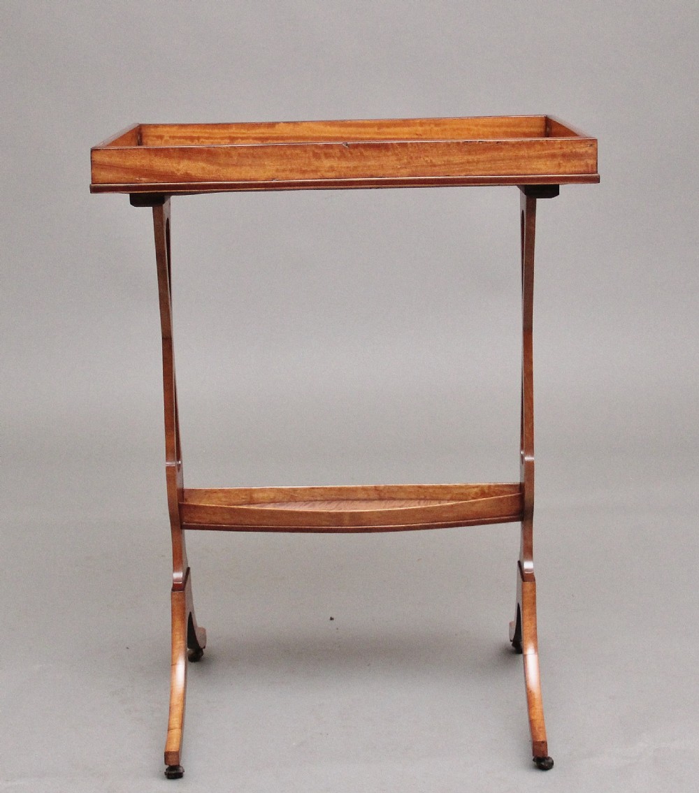 A 19th-century Sheraton Revival satinwood serving table featuring a tray top, pierced side supports, a lower shelf, and splayed legs on brass castors. View X