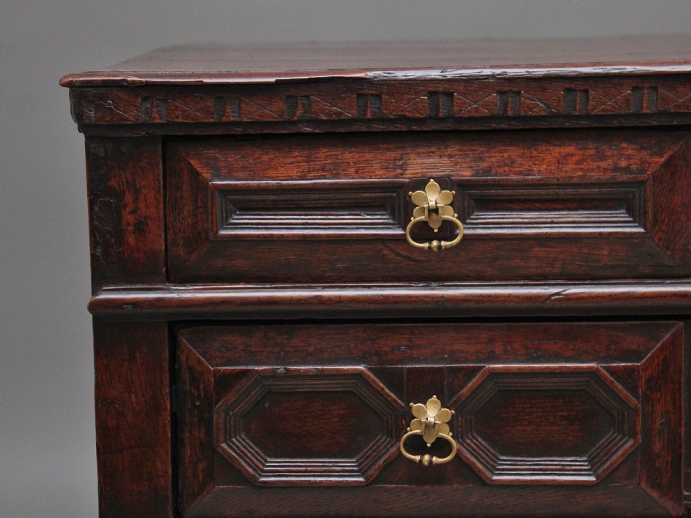 An early eighteenth century dark oak chest of drawers featuring geometric moulded drawer fronts, carved frieze, and brass drop handles. View II