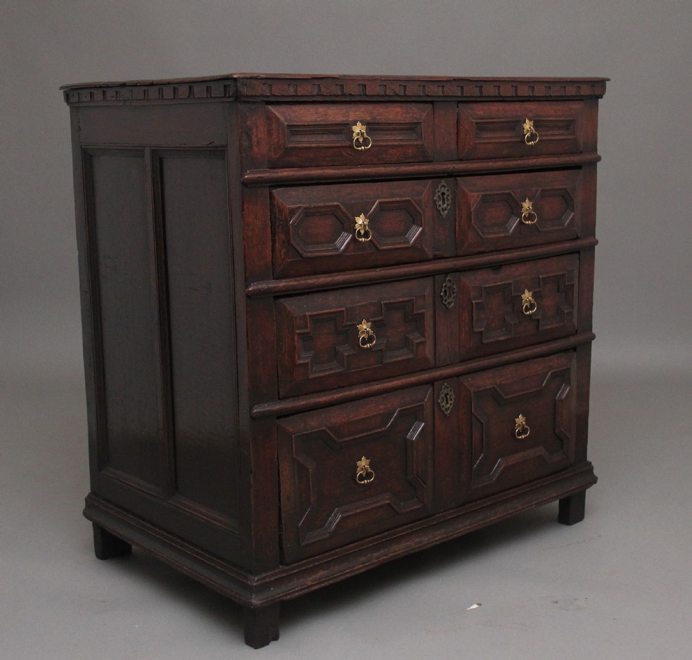 An early eighteenth century dark oak chest of drawers featuring geometric moulded drawer fronts, carved frieze, and brass drop handles. View V