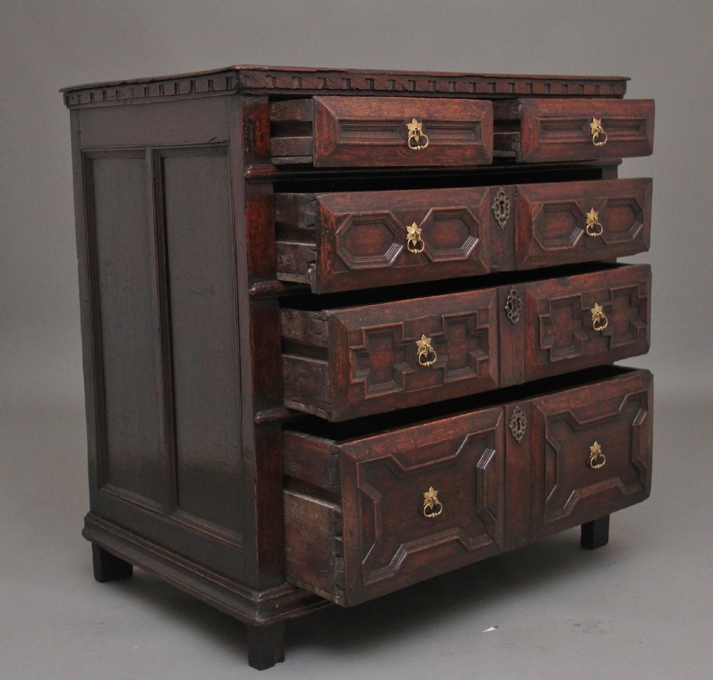 An early eighteenth century dark oak chest of drawers featuring geometric moulded drawer fronts, carved frieze, and brass drop handles. View VI