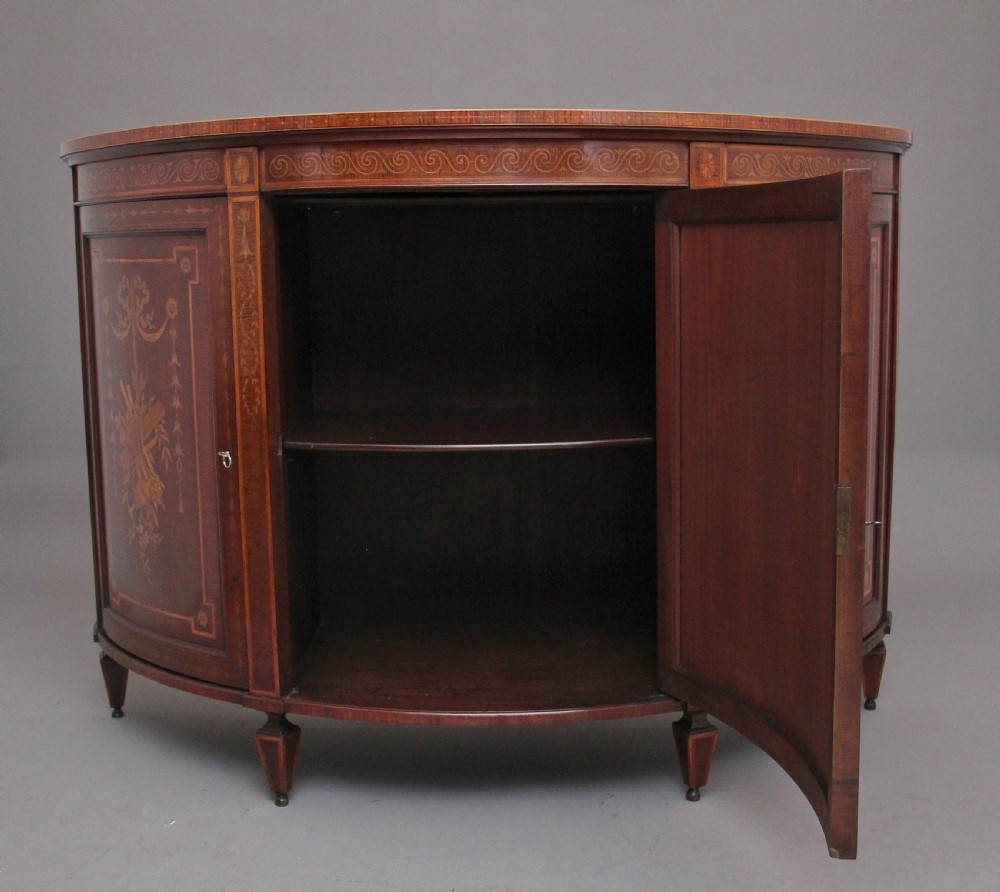 A 19th-century curved mahogany cabinet featuring intricate satinwood marquetry panels depicting classical figures, swags, and scrollwork. View IV