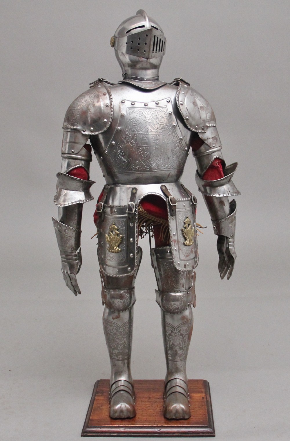 A detailed miniature suit of polished steel plate armour with fine surface etchings, brass eagle emblems, and red fabric lining, standing on a dark wood base.
