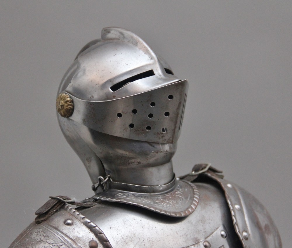 A detailed miniature suit of polished steel plate armour with fine surface etchings, brass eagle emblems, and red fabric lining, standing on a dark wood base. View IX