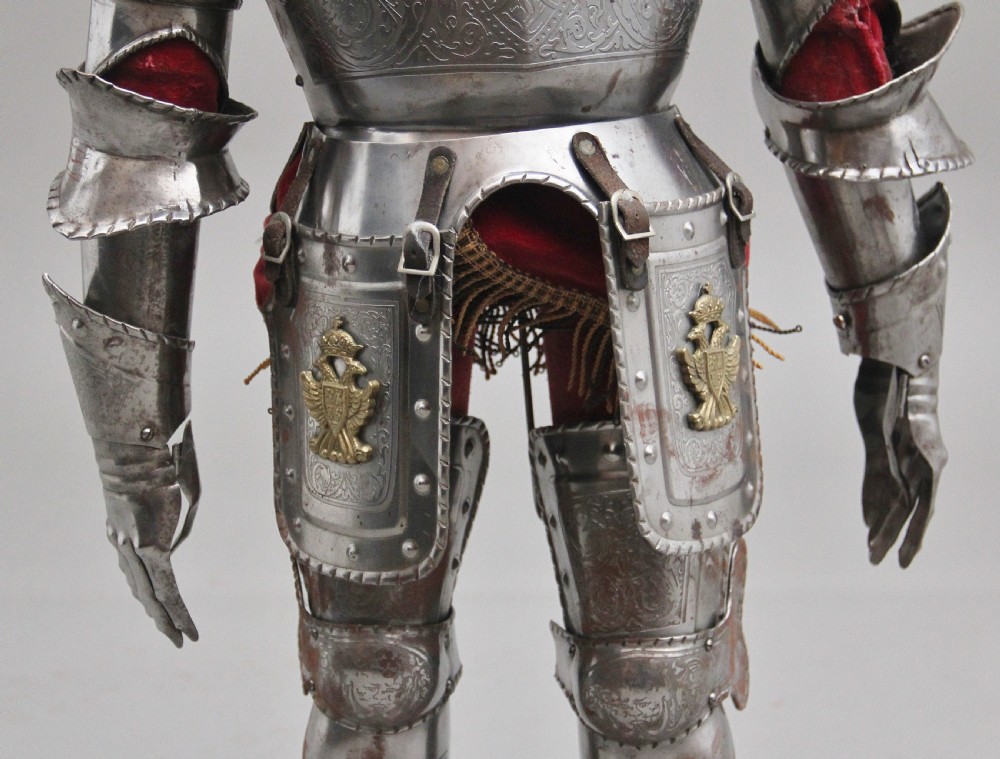 A detailed miniature suit of polished steel plate armour with fine surface etchings, brass eagle emblems, and red fabric lining, standing on a dark wood base. View XI