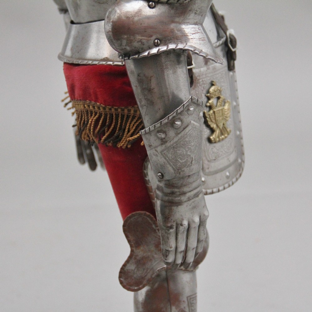 A detailed miniature suit of polished steel plate armour with fine surface etchings, brass eagle emblems, and red fabric lining, standing on a dark wood base. View XII