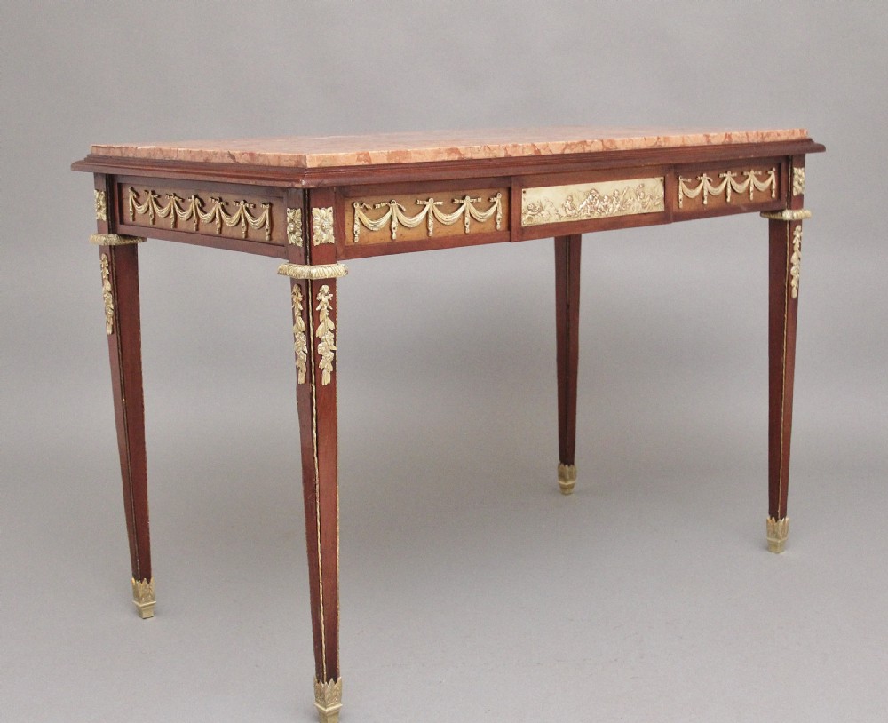 A 19th-century French mahogany centre table featuring a pink marble top, slender tapered legs, and decorative gilt-bronze mounts with swags and cherub relief. View VI