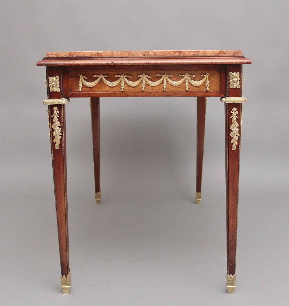 A 19th-century French mahogany centre table featuring a pink marble top, slender tapered legs, and decorative gilt-bronze mounts with swags and cherub relief. View X