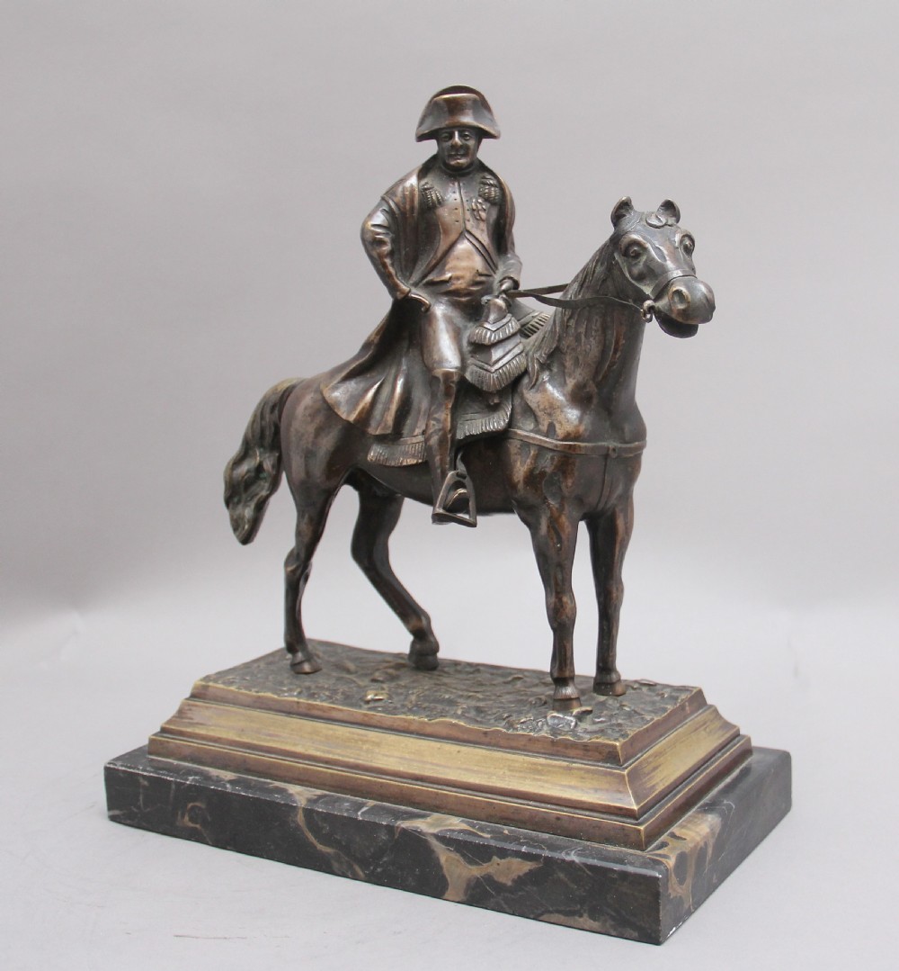 A dark bronze equestrian sculpture depicting Napoleon Bonaparte on horseback mounted on a stepped brass and black marble plinth.