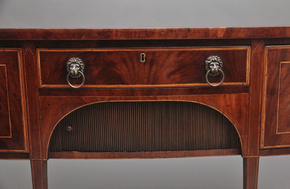 An early nineteenth-century mahogany bowfront sideboard with line inlay, central arch detail, brass lion mask ring handles, and tapered square legs. View VI