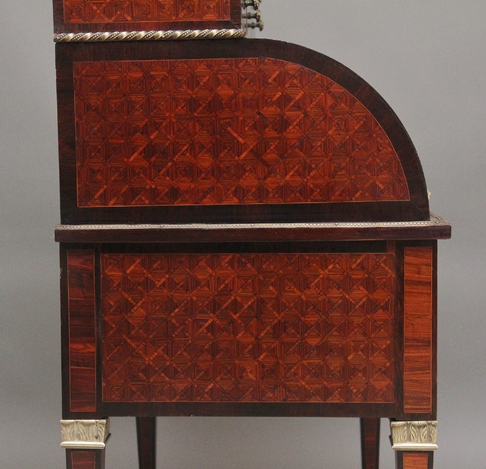 A 19th-century French kingwood cylinder desk featuring parquetry veneering, gilt-brass mounts, tapered square legs, and a curved roll-top section over multiple small drawers. View XVIII