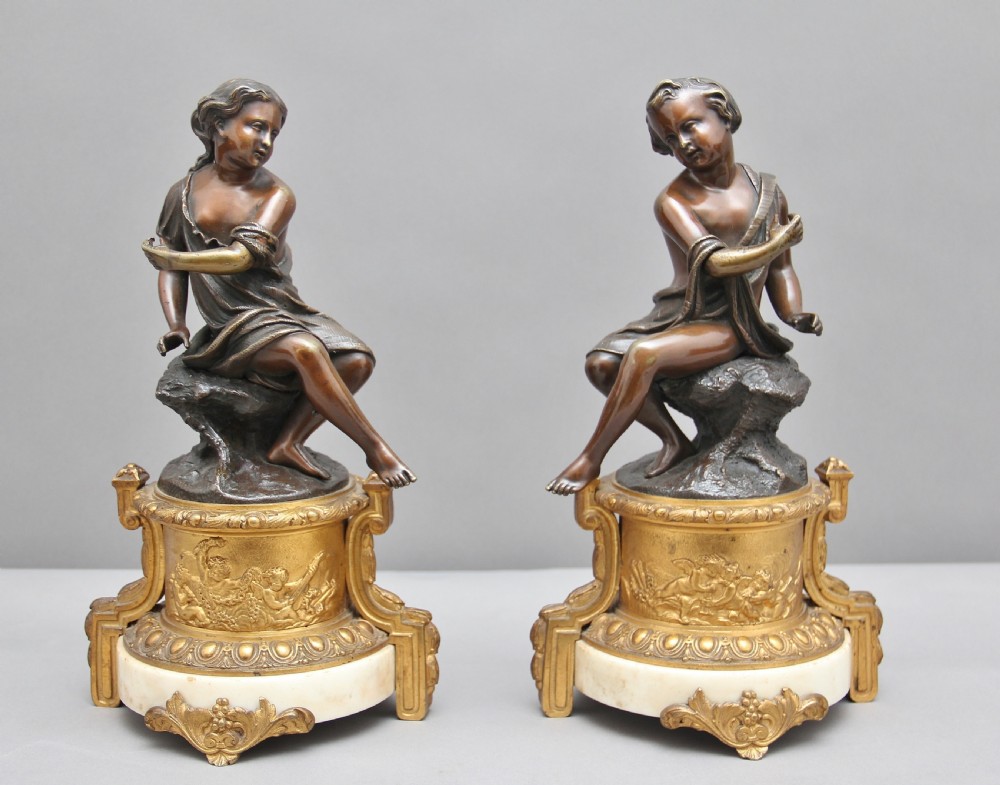 A pair of patinated dark bronze figurative statuettes depicting classical youths seated on rocky outcrops, set atop ornate gilded ormolu and white marble bases.