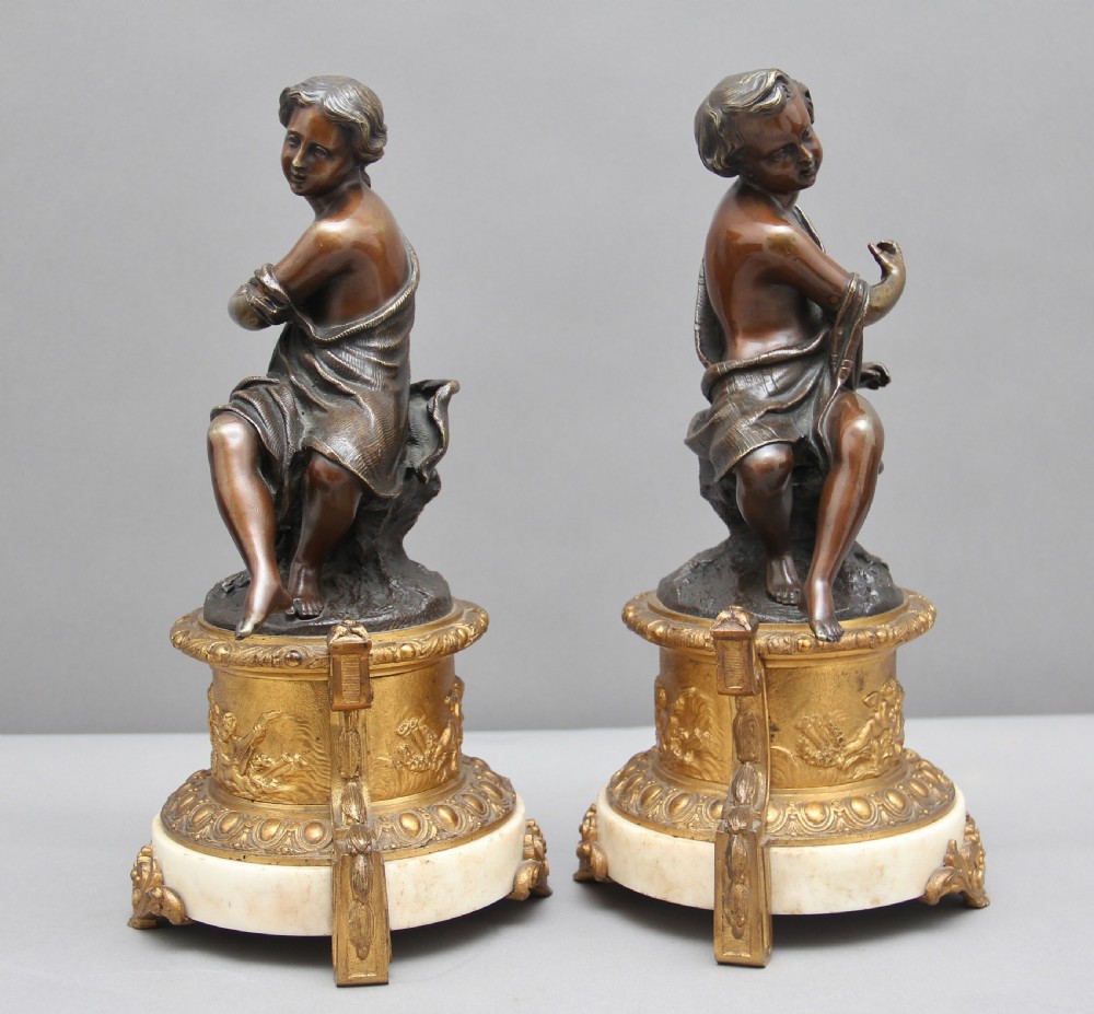 A pair of patinated dark bronze figurative statuettes depicting classical youths seated on rocky outcrops, set atop ornate gilded ormolu and white marble bases. View II