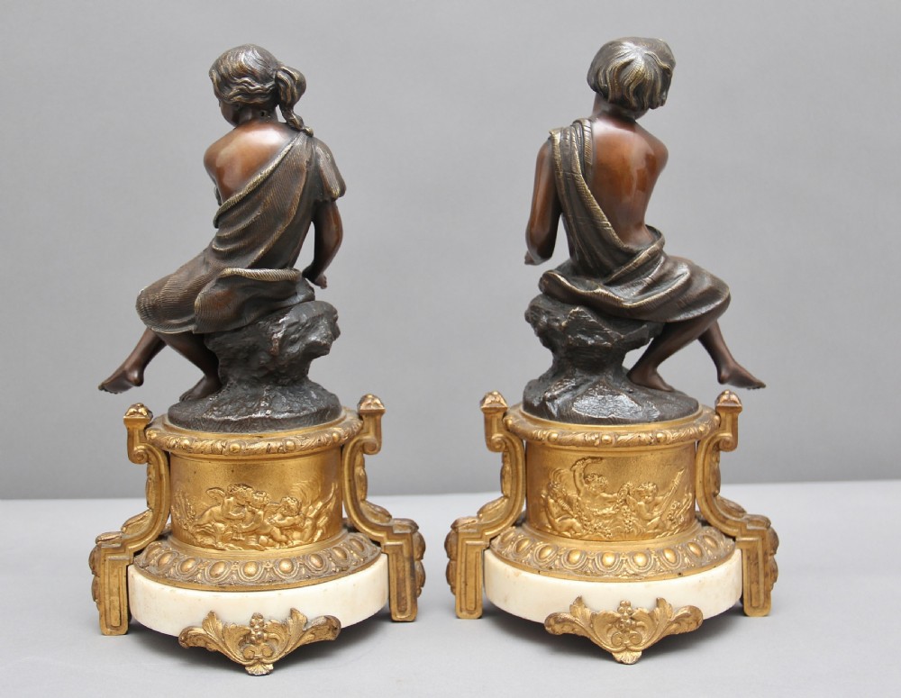 A pair of patinated dark bronze figurative statuettes depicting classical youths seated on rocky outcrops, set atop ornate gilded ormolu and white marble bases. View III