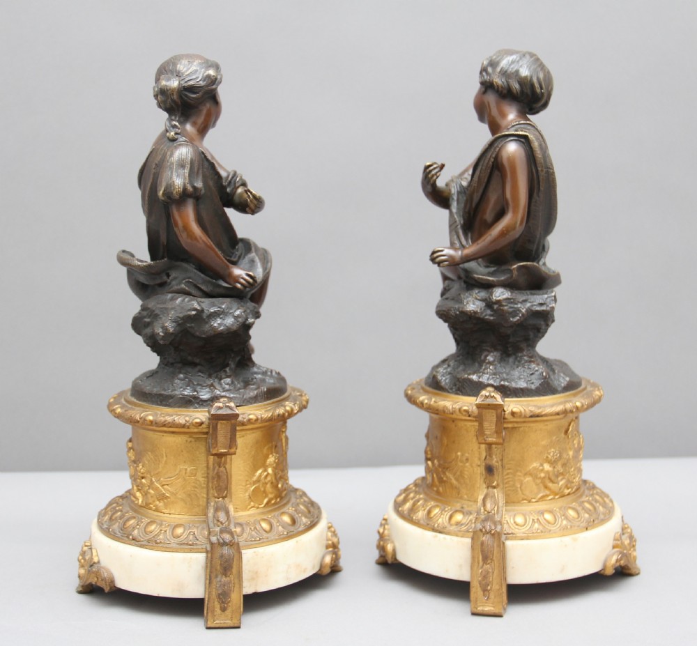 A pair of patinated dark bronze figurative statuettes depicting classical youths seated on rocky outcrops, set atop ornate gilded ormolu and white marble bases. View V