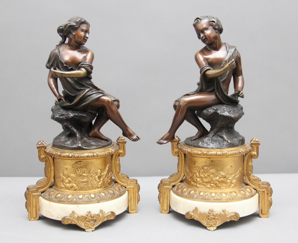 A pair of patinated dark bronze figurative statuettes depicting classical youths seated on rocky outcrops, set atop ornate gilded ormolu and white marble bases. View VI