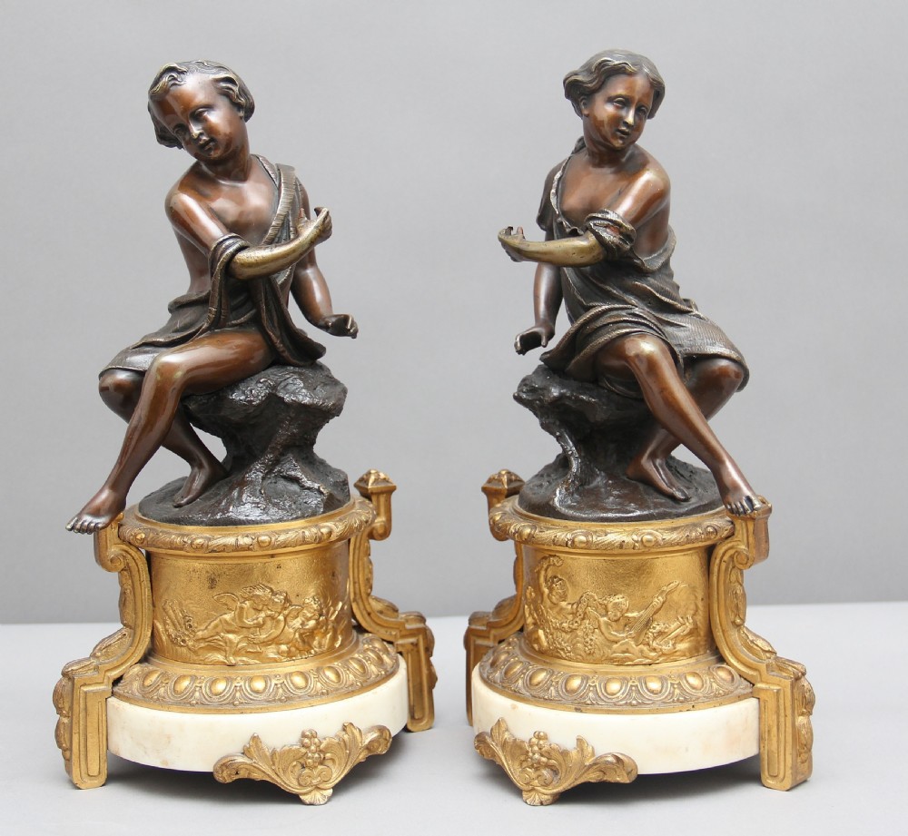 A pair of patinated dark bronze figurative statuettes depicting classical youths seated on rocky outcrops, set atop ornate gilded ormolu and white marble bases. View VII
