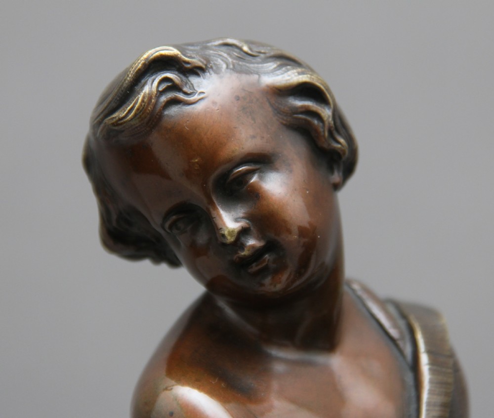 A pair of patinated dark bronze figurative statuettes depicting classical youths seated on rocky outcrops, set atop ornate gilded ormolu and white marble bases. View VIII