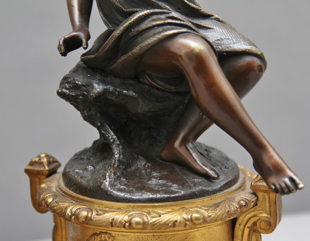A pair of patinated dark bronze figurative statuettes depicting classical youths seated on rocky outcrops, set atop ornate gilded ormolu and white marble bases. View X