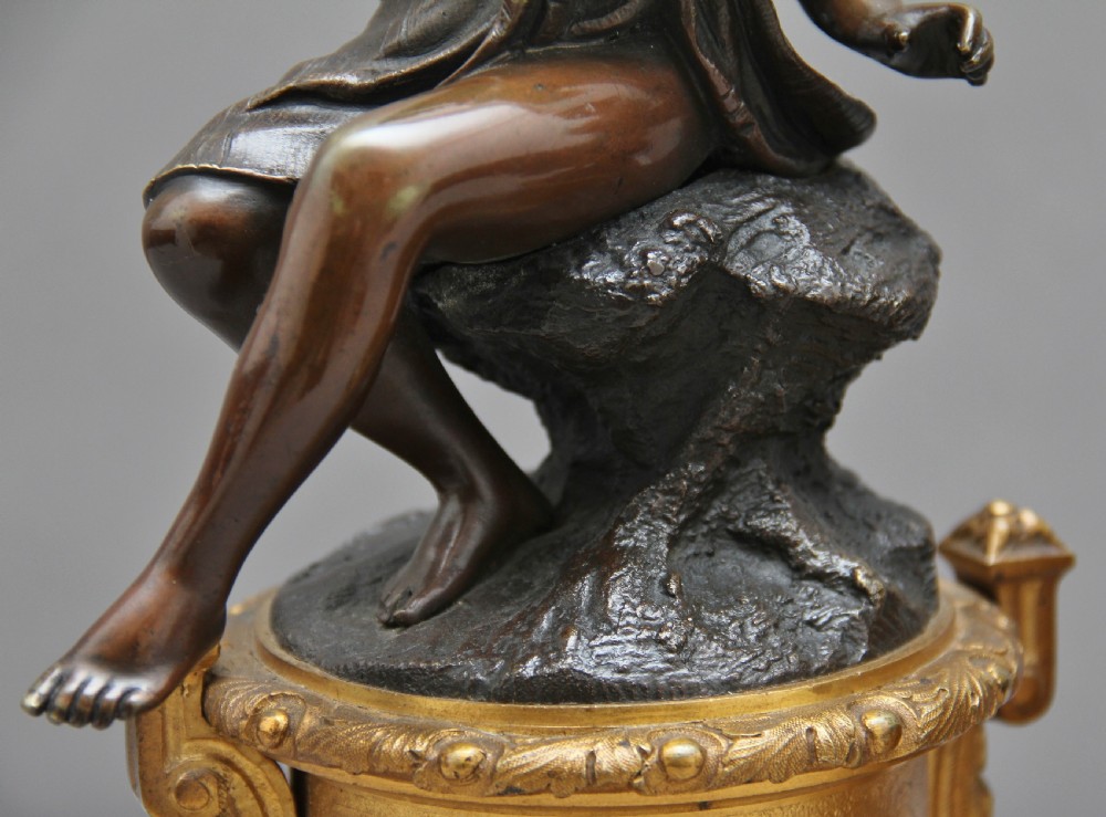 A pair of patinated dark bronze figurative statuettes depicting classical youths seated on rocky outcrops, set atop ornate gilded ormolu and white marble bases. View XI